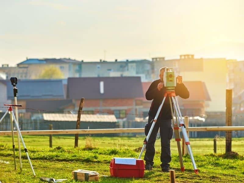 Land surveyor measuring former orchard property lines beside modern residential development.